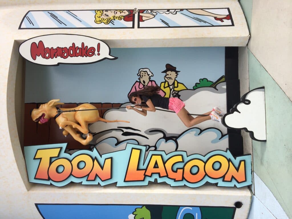 Foto que mostra jovem adulta pendurada na coleira do cachorro que está correndo com um balão de fala "Marmaduke"que é o nome do cachorro. Ao fundo há um casal de senhores que estão surpresos com a cena e na frente o letreiro TOON LAGOON, nome da área temática fo parque