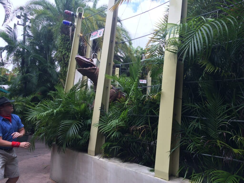 Foto que mostra um velociraptor, dinossauro do filme Jurassic World, com com a boca aberta rugindo junto a seu treinador, que recua ligeiramente assustado com a atitude do dinossauro.
