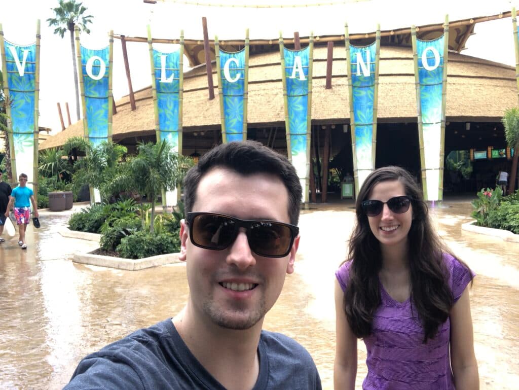 Casal jovem em frente a entrada do parque aquático Volcano Bay, na Universal Studios Orlando. Ambos usam óculos de sol e nos fundos tem o letreiro escrito VOLCANO