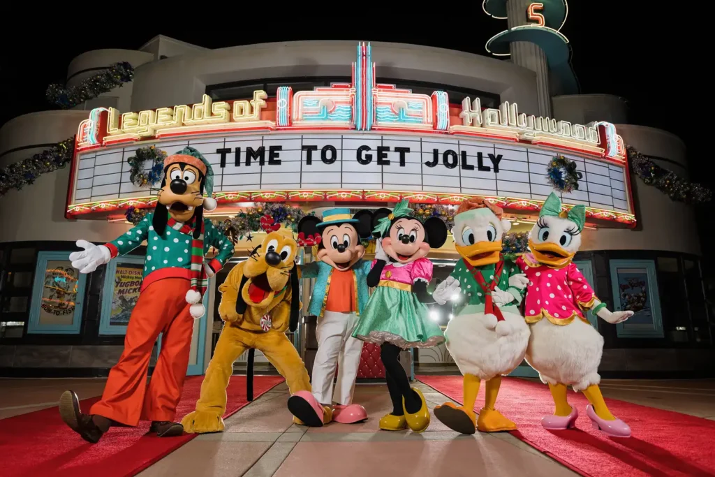 Personagens da Disney posam animados em frente a um letreiro retrô iluminado que diz "Legends of Hollywood, Time to Jolly". Os personagens Mickey Mouse, Minnie Mouse, Pluto, Pato Donald, Margarida e Pateta estão no parque Hollywood Studios com roupas festivas de natal em estilo retrô elegante