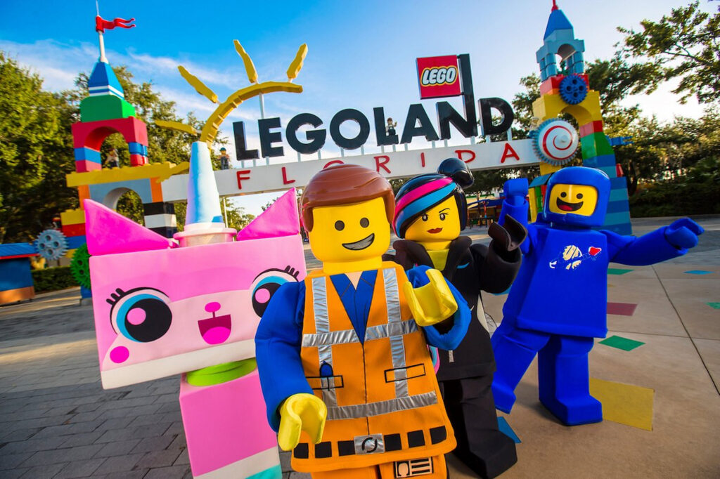 Personagens feitos de lego cumprimentando em frente a entrada do parque, também toda construída de lego, que diz Legoland Florida, tem uma pequena bandeira vermelha escrito Lego, e um sol do outro lado