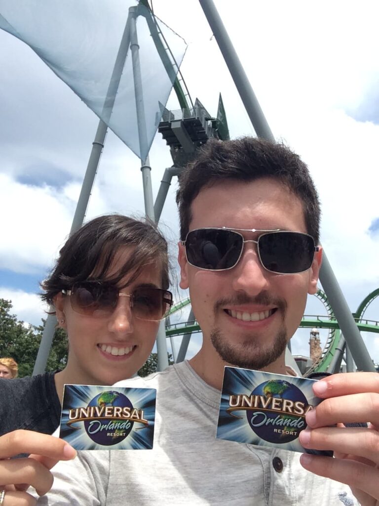 Casal se jovens usando óculos de sol sorri mostrando seus passes de fura filas da Universal Studios Orlando. Nessa passe é possível ver o logo da Universal com seu globo e ao fundo dá também para ver a montanha russa do Hulk