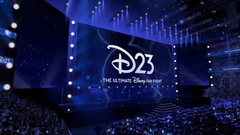 Palco com telão enorme ao fundo, mostrando D23 escrito e totalmente iluminado nas cores do evento que são tons de azul, branco e prata. Pessoas sentadas na platéia aguardando ansiosamente pelo início do evento