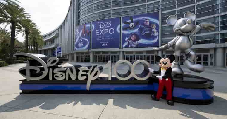 Foto do Mickey Mouse em frente ao Anaheim Convention Center, local onde ocorre o evento D23 a cada dois anos. Mickey está na Frente de Um letreiro escrito Disney100, referente a comemoração de 100 anos da Disney que está junto a uma estátua prata também do Mickey. Mais atrás há um cartaz da D23 Expo