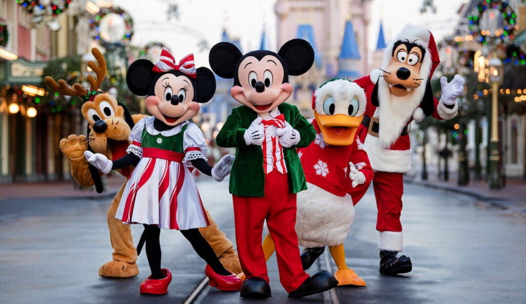 Foto mostra Mickey, Minnie, Donald, Pluto e Pateta em trajes natalinos na Main Street USA, na entrada do Parque de diversões Magic Kingdom, em Walt Disney World Resort, na Flórida. O parque está decorado para o natal e o Castelo da Cinderela encontra-se ao fundo. Pateta usa uma fantasia de papai noel, enquanto Puto está com orelhas de Rena
