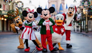 Foto mostra Mickey, Minnie, Donald, Pluto e Pateta em trajes natalinos na Main Street USA, na entrada do Parque de diversões Magic Kingdom, em Walt Disney World Resort, na Flórida. O parque está decorado para o natal e o Castelo da Cinderela encontra-se ao fundo. Pateta usa uma fantasia de papai noel, enquanto Puto está com orelhas de Rena