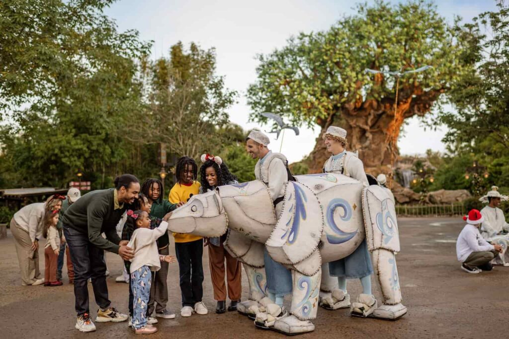 Na entrada do Animal Kingdom, em frente a Arvore da Vida, dois homens estão dentro de uma estrutura em formato de Urso Polar articulado, que os permite interagirem com os visitantes do parque, como fazem na foto, onde temos três crianças e um adolescente acariciando o urso, junto de seus pais