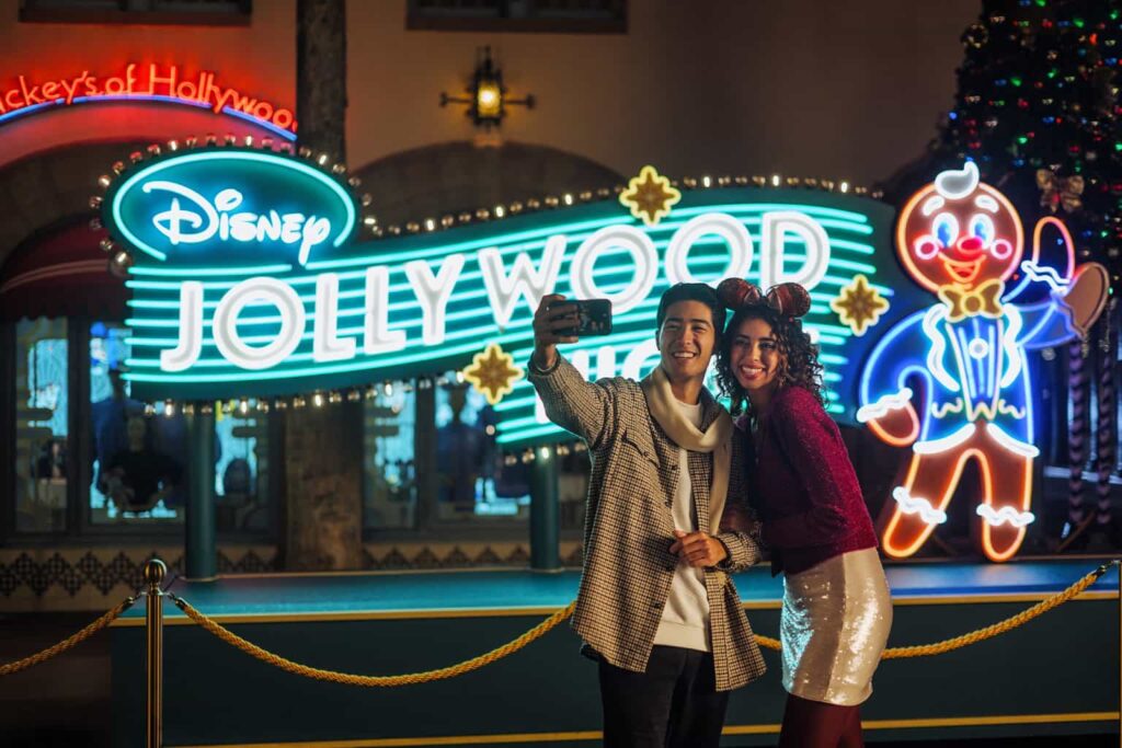 Casal bem vestido tira foto em frente ao letreiro da festa Jollywood Nights, a festa de natal do parque da Disney Hollywood Studios, que conta com um biscoito de natal ao lado das letras