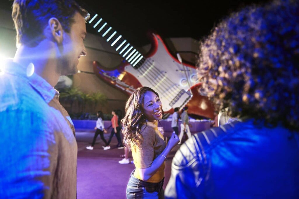 Em frente a guitarra da atração Rock'n'Roller Coaster, no parque de diversões Hollywood Studios em Walt Disney World Resort, uma muher mais a frente, está andando em direção a atração enquanto seu amigos um pouco mais atrás a acompanham.