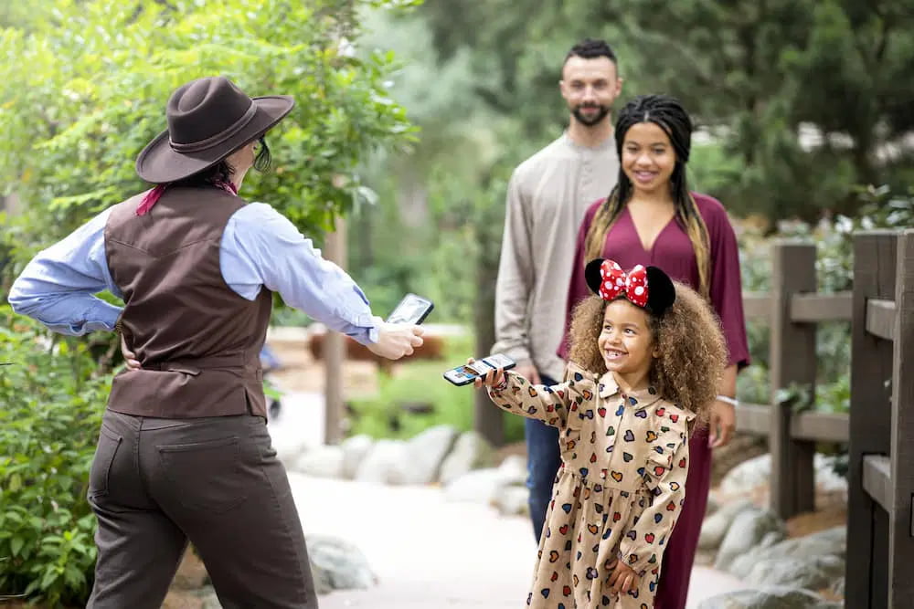 Cast member escaneia celular na mão de uma garotinha que usa orelhinhas da minnie e está sorrindo ao mostrar seu Lightning Lane Premier Pass, que vai possibilitar que ela e seus pais, que estão logo atrás, furem a fila da atração. Os pais ao fundo sorriem ao ver a cena