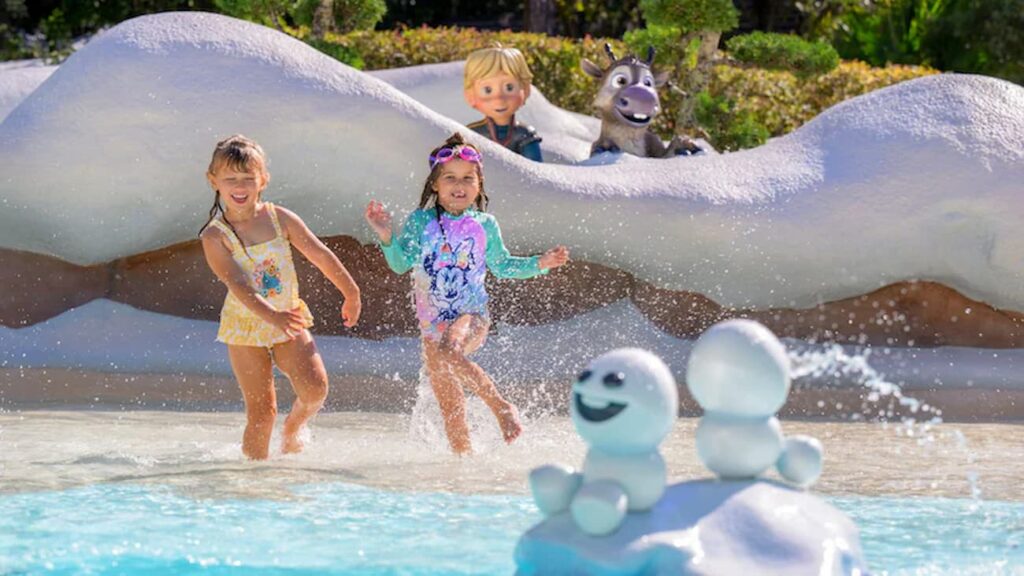 Meninas brincam no parque aquático Disney's Blizzard Beach, na área infantil do parque. Ambas estão na água rasa, a frente temos pequenos bonecos de neve do filme Frozen e atrás, temos o Kristoff ainda criança, acompanhado do pequeno Sven, seu fiel amigo