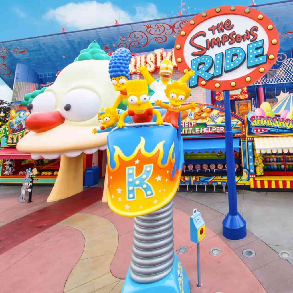 foto da entrada da atração The Simpsons Ride, no parque Universal Studios Orlando, mostra Homer, Marge, Bart, Lisa e Maggie, em um carrinho de montanha russa, ao lado do nome da atração e também de uma cabeça gigante do palhaço Krusty, com a boca aberta, entrada do simulador