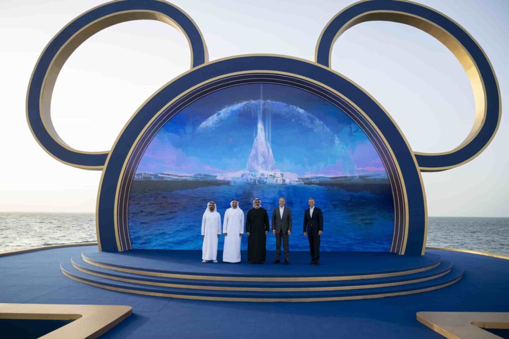 Mohamed Abdalla Al Zaabi (CEO da Miral), Sua Excelência Mohamed Khalifa Al Mubarak (Presidente da Miral), Sua Alteza Sheikh Khaled bin Mohamed bin Zayed Al Nahyan (Príncipe Herdeiro de Abu Dhabi), Bob Iger (CEO da The Walt Disney Company) e Josh D’Amaro (Presidente da Disney Experiences). Todos estão em frente uma moldura em formato de Mickey que dentro possui a imagem ilustrativa conceitual do projeto anunciado