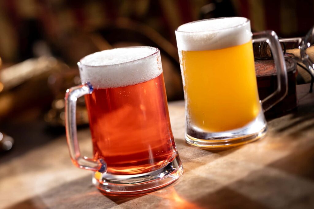 duas canecas lado a lado, com bebidas que parecem cervejas, uma mais tradicional e outra num tom mais alaranjada, ambas em uma mesa num cenário meio pirata no estilo do novo bar Beak and Barrel que inaugura no Magic Kingdom em Agosto de 2025