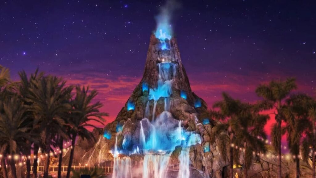 Imagem ilustrativa do vulcão do parque aquático temático Volcano Bay. O vulção está em frente um entardecer já quase a noite, completamente iluminado, fazendo propaganda para o Evento Volcano Bay Nights que acontece em noites selecionados no verão americano, tem ingresso a parte e muita diversão