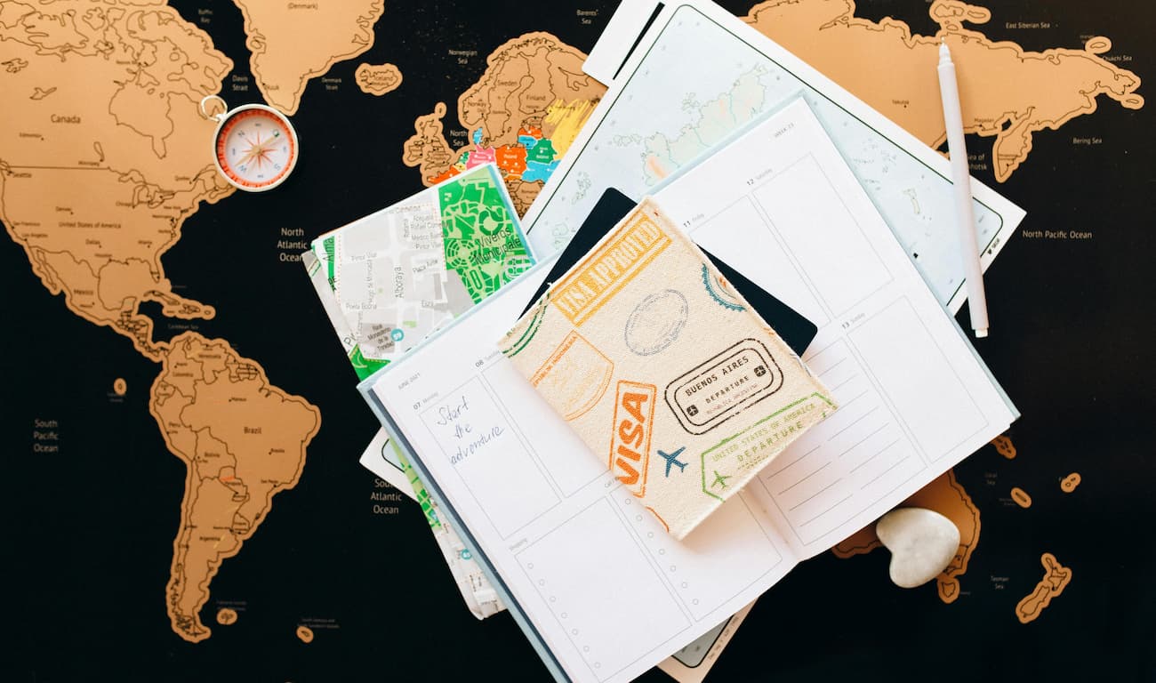 cadernos e documentos de viagem, incluindo um passaporte com a estampa de carimbos de passaporte com as letras USA referindo-se aos estados unidos em cima do mapa mundi indicando planejamento de viagem para os estados unidos, onde é necessários visto de turismo para brasileiros