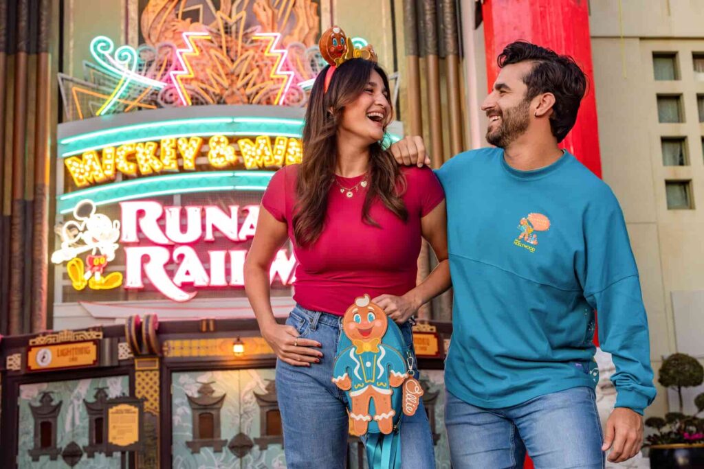 Casal posa olhando um para o outro e sorrindo com seus acessórios adquiridos na festa de natal Jollywood Nights do Hollyood Studios em Walt Disney World Resort. É possível vê-los usando Spirit Jersey e mochila da Loungefly, todos com o simbolo da festa que é um gingerbread de terninho