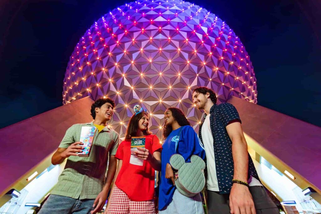 Quatro amigos, dois homens e duas mulheres jovens, em frente a Spaceship Earth, simbolo do parque Epcot, iluminada a. noite, parecem se divertir enquanto estão no parque, um deles tem um pote de pipoca na mão e a outra um mapa do paruqe