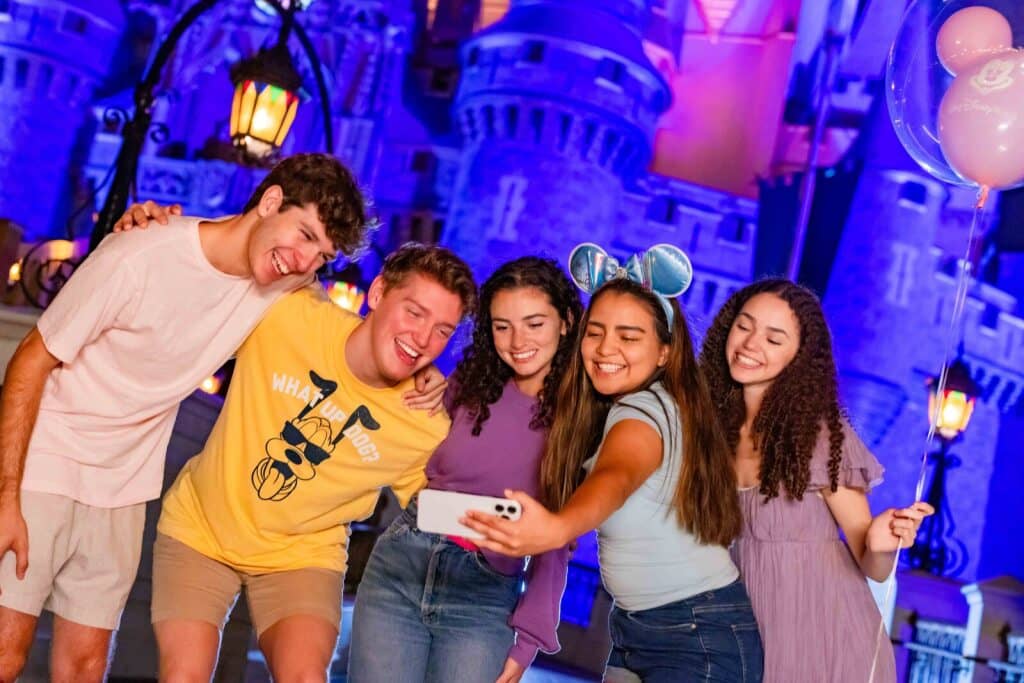 2 homens jovens posam para foto se abraçando junto com mais 3 mulheres também jovens, sendo que uma delas captura o momento em uma selfie com seu celular em frente ao Castelo da Cinderela, a noite, no Magic Kingdom em Walt Disney World Resort