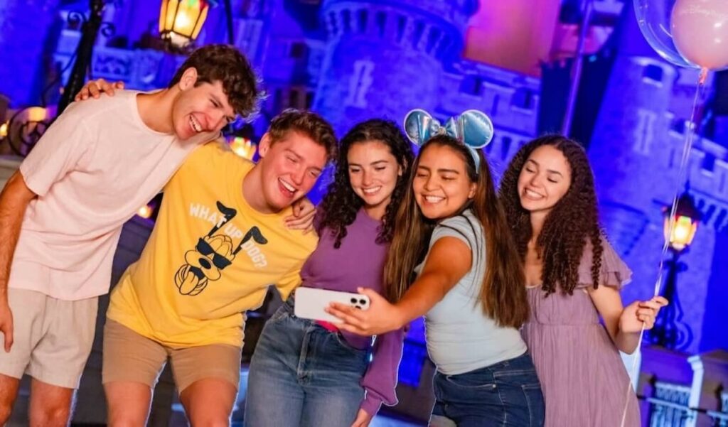 2 homens jovens posam para foto se abraçando junto com mais 3 mulheres também jovens, sendo que uma delas captura o momento em uma selfie com seu celular em frente ao Castelo da Cinderela, a noite, no Magic Kingdom em Walt Disney World Resort