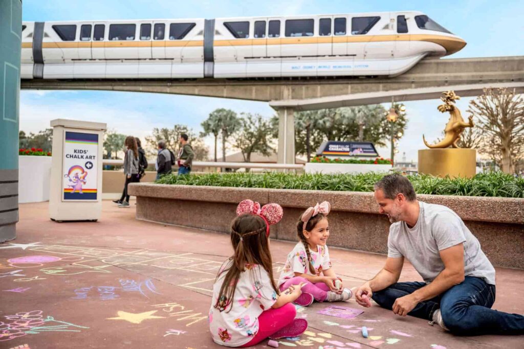 Homem brinca com duas meninas crianças que usam orelhinhas da Minnie Mouse, de pintar com giz de cera no chão, dentro do parque Epcot, com o Monorail passando ao fundo, durante o International Festival of the Arts de Walt Disney World Resort
