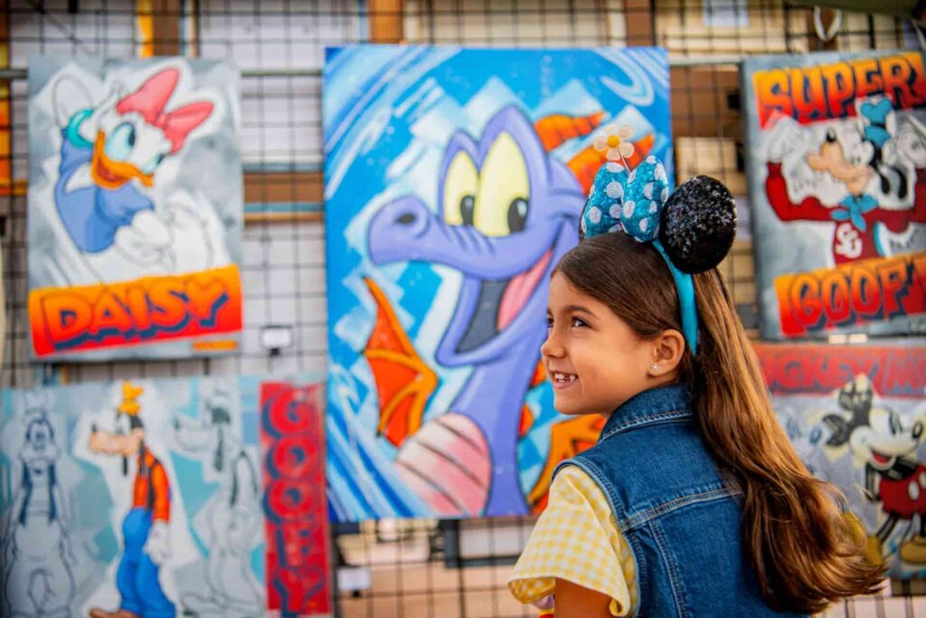 Menininha usando orelhinhas da Minnie em frente a um quadro do Figment, simbolo do parque Epcot, que recebe anualmente o Festival of the Arts, festival que celebra a arte em Orlando