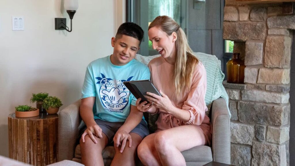 Mãe e filho sentados confortavelmente em uma grande poltrona com tablet na mão participam de uma conversa por vídeo para ter acesso ao DAS na Disney