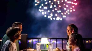 família sentada a mesa come enquanto assistem ao show de fogos do parque da Disney, o Epcot