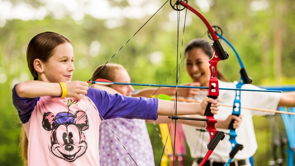 Menina com camiseta da Minnie usando arco e flecha em destaque e mais ao fundo uma mulher ensina uma outra pessoa a fazer o mesmo, todos se divertem bastante na atividade que acontece no Fort Wilderness Resort, Hotel em Walt Disney World