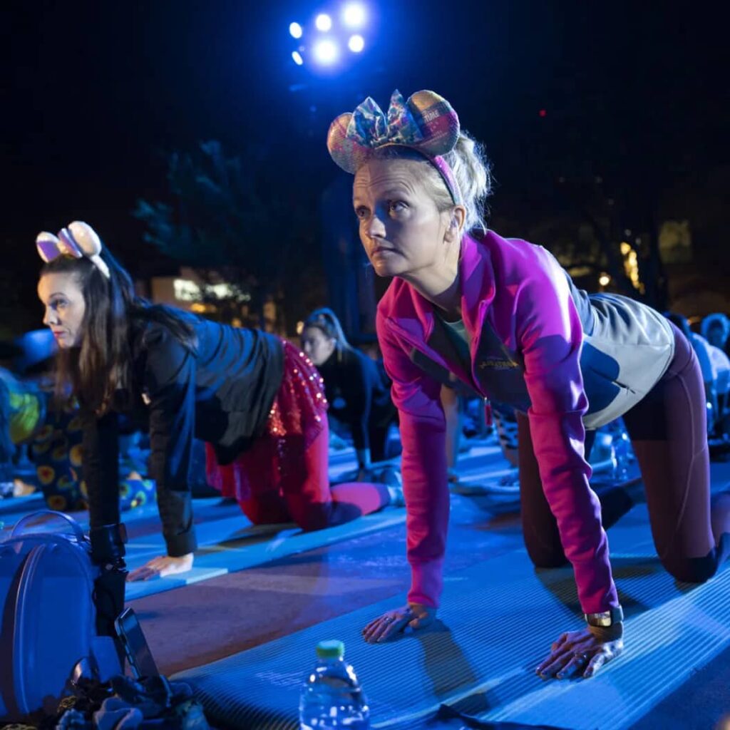 Mulheres com roupas de ginástica sentadas em tapete de de yoga e usando orelhinhas da Minnie fazem posição de yoga