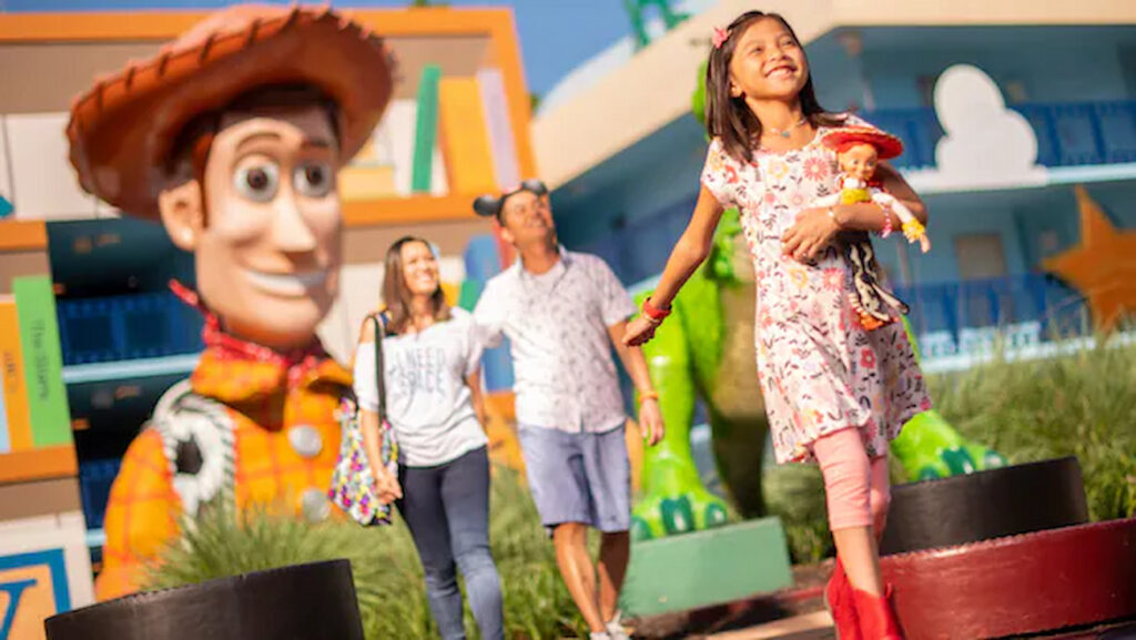 Menina segura uma boneca da Jessie de Toy Story enquanto anda com seus pais, meio aos prédios do Hotel Disney's All Star Movies em Orlando no complexo Walt Disney World Resort