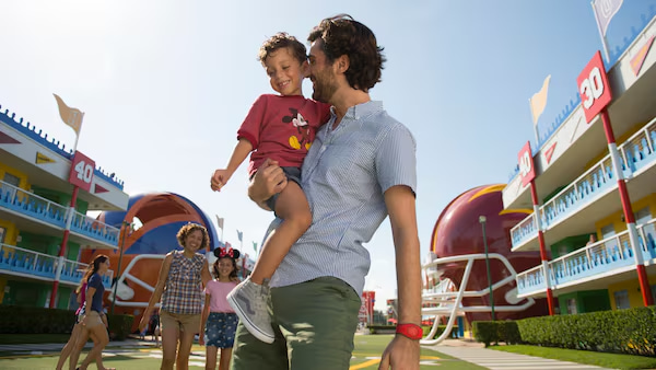 Pai com seu filho no colo, a frente, e mais atrás mãe vem acompanhando com sua filha, meio aos prédios do Hotel Disney's All Star Sports em Orlando no complexo Walt Disney World Resort