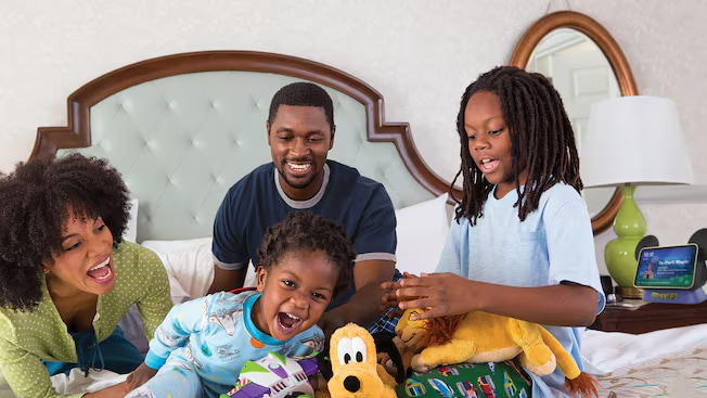 Família se diverte na cama de hotel Disney com a Alexa Hey Disney ao lado junto de uma pelúcia do Pluto