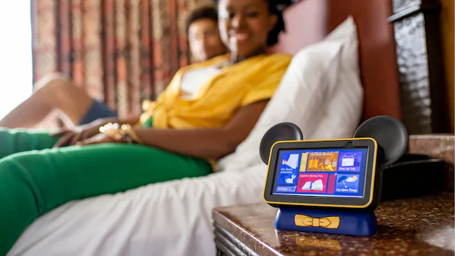 Casal sentado na cama de hotel Disney observa a Hey Disney que é a Alexa personalizada especialmente para o resort dos hotéis da Disney