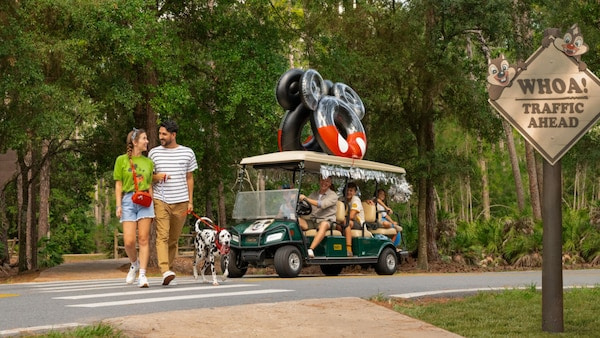 Casal com cachorro anda pelo hotel Fort Wilderness Resort, enquanto família vem em um carrinho de golfe mais atrás, meio de transporte muito comum dentro desse hotel que é muito grande no complexo de Walt Disney World Resort
