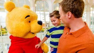 Pai segura seu filho no colo enquanto Pooh interage com a criança no restaurante Crystal Palace onde é possível encontrar o Ursinho Pooh e seus amigos durante refeições no café da manhã, almoço e jantar