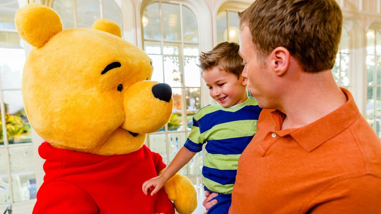 Pai segura seu filho no colo enquanto Pooh interage com a criança no restaurante Crystal Palace onde é possível encontrar o Ursinho Pooh e seus amigos durante refeições no café da manhã, almoço e jantar