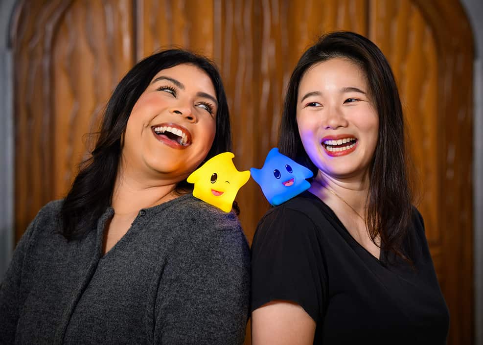 Mulheres se olham e sorriem com estreilinhas brilhantes - shoulder pals - em seus ombros, que são do merchandising especial da comemoração do dia do mário