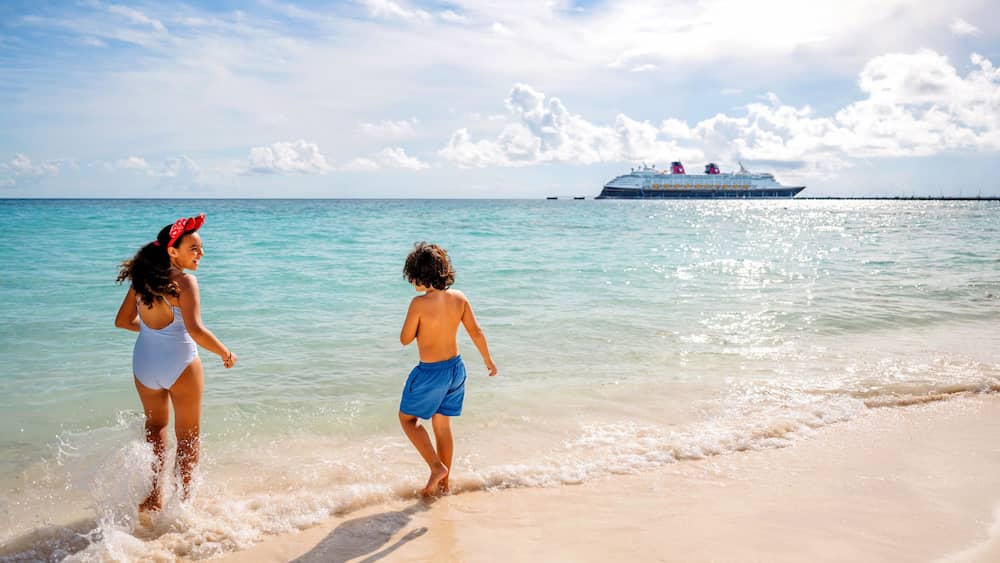 Crianças brincam na beira da praia com água cristalina e navio da Disney ao fundo