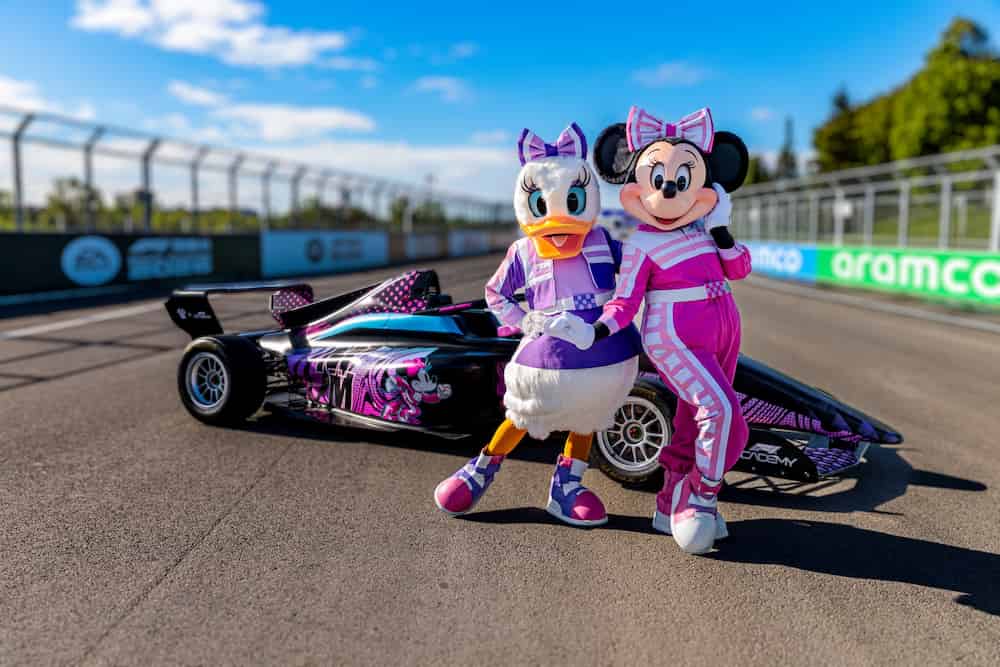 Minnie e Margarida vestidas com roupas de corredoras de fórmula 1 posam ao lado de carro temático numa pista de corrida