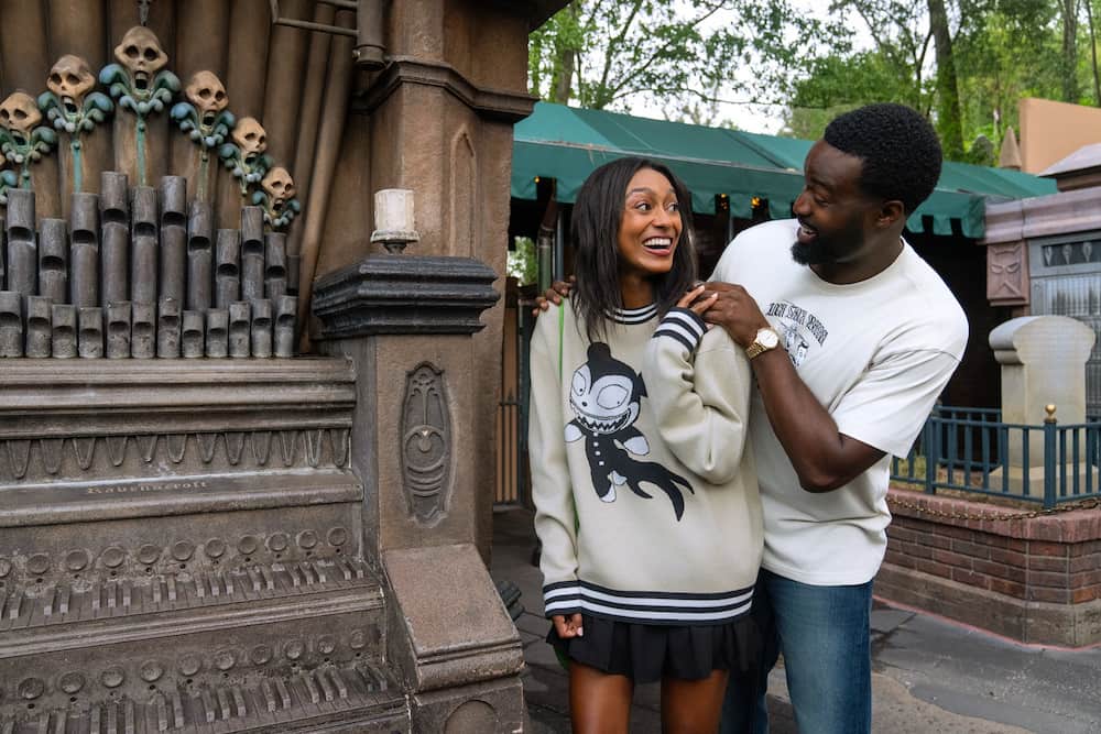 Casal posa na fila na fila da atração da mansão assombrada enquanto usa roupas da coleção Haunted Mansion