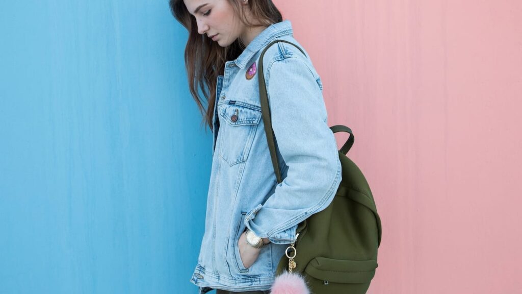 Menina de cabeça baixa e jaqueta jeans carrega sua mochila em frente a uma parede metade rosa e metade azul