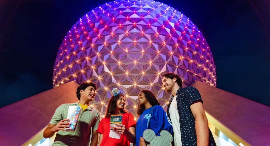 Quatro amigos, dois homens e duas mulheres jovens, em frente a Spaceship Earth, simbolo do parque Epcot, iluminada a. noite, parecem se divertir enquanto estão no parque, um deles tem um pote de pipoca na mão e a outra um mapa do parque