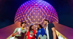Quatro amigos, dois homens e duas mulheres jovens, em frente a Spaceship Earth, simbolo do parque Epcot, iluminada a. noite, parecem se divertir enquanto estão no parque, um deles tem um pote de pipoca na mão e a outra um mapa do parque