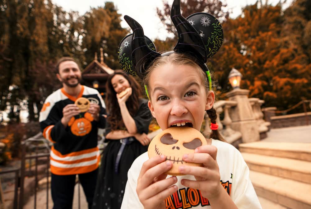 Menina com orelhinhas da Minnie Malevola come seu cookie do Jack de Estranho mundo de Jack mais a frente enquanto seus pais ao fundo fazem o mesmo durante o Halloween da Disneyland Paris