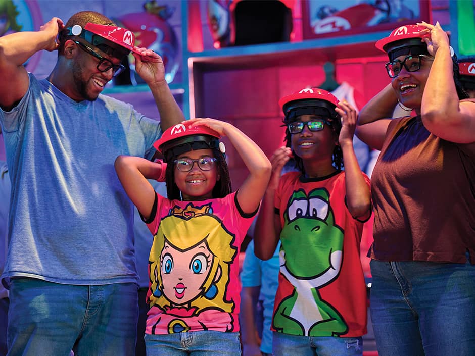 Familia com mãe, pai, filho e filha se preparam para entrar na atração Mario Kart colocando seus chapéus onde são acoplados óculos interativos para pontuar